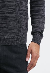 Mikina s kapucí v antracitové šedé barvě s proužky, s přední kapsou a žebrovanými manžetami. Materiál má texturu a kombinuje tmavší a světlejší odstíny.