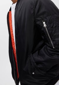 Veste bomber noire avec doublure en tissu matelassé orange, poches latérales et poche zippée sur la manche. Texture lisse avec col côtelé.
