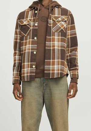 Mand iført brun ternet skjorte over brun hættetrøje og slidte blå jeans, stående foran en ensfarvet baggrund.