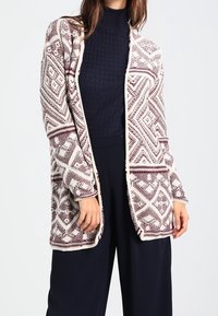 Cardigan a maglia con un motivo geometrico bordeaux e crema, davanti aperto, maniche lunghe e una vestibilità rilassata. Indossato sopra un maglione scuro e pantaloni.