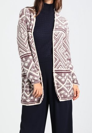 Cardigan a maglia con un motivo geometrico bordeaux e crema, davanti aperto, maniche lunghe e una vestibilità rilassata. Indossato sopra un maglione scuro e pantaloni.