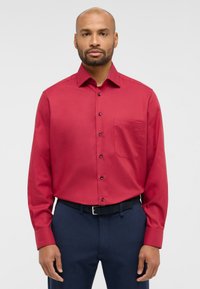 Camicia rossa a maniche lunghe con bottoni e taschino sul petto. Presenta un colletto e bottoni neri, abbinata a pantaloni blu scuro e una cintura nera.