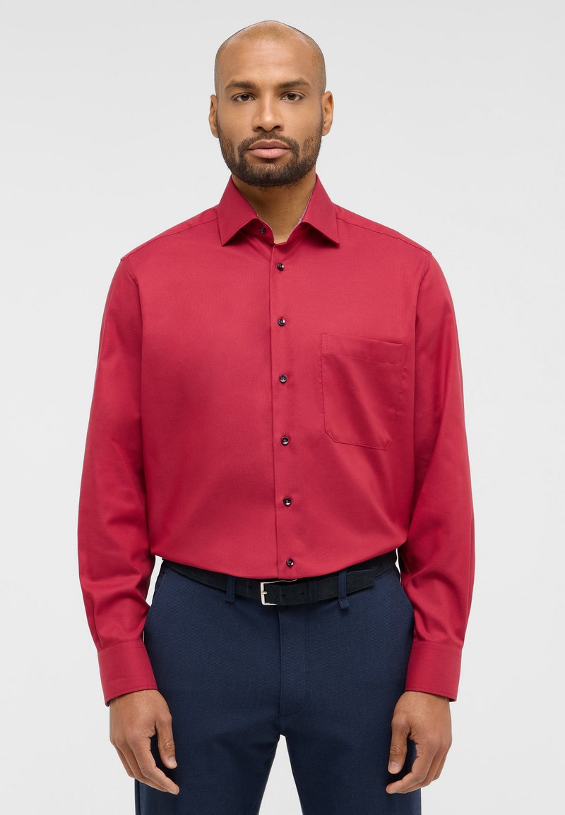 Camicia rossa a maniche lunghe con bottoni e taschino sul petto. Presenta un colletto e bottoni neri, abbinata a pantaloni blu scuro e una cintura nera.