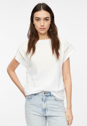 Woman with long brown hair wearing a white short-sleeve top and light blue denim jeans, standing with one hand on hip.