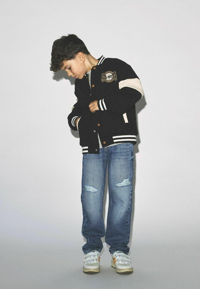 Veste varsity noire avec des manches crème et des poignets rayés, dotée d'un patch logo. Portée avec un jean bleu déchiré et des baskets blanches.