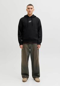 Hoodie en coton noir avec impression du logo, cordons de serrage, poche kangourou, assorti à un pantalon beige oversized et des chaussures de couleur claire.