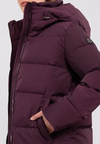 Pflaumenfarbene Steppjacke mit Kapuze, ausgestattet mit einem Reißverschluss, seitlichen Taschen und einem runden Logo-Akzent am Ärmel. Glatte Oberfläche.