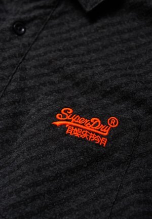 Zbliżenie ciemnej, teksturowanej tkaniny z jasnopomarańczowym, haftowanym logo "SuperDry" oraz małym wszytym guzikiem powyżej.