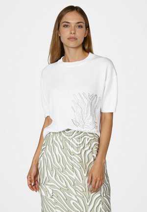 Frau mit langem braunem Haar, die einen weißen kurzärmeligen Pullover mit Strass-Design und einen grün-weiß gemusterten Rock trägt.