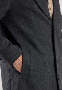 Cappotto di lana grigio scuro con colletto a revers, tasche laterali e una superficie liscia, con chiusura a bottoni visibile e vestibilità su misura.