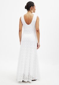 Witte kanten maxi-jurk met een ronde rug en een aansluitende silhouette, met een uitlopende onderkant en bloemmotief. Geddragen met grote stervormige oorbellen.