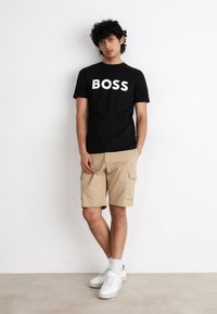 Melnā T-kreklā ar baltu uzrakstu "BOSS", savienots ar beigu krāsu krājuma šortiem. Modelis valkā baltus apavus un potītes garuma zeķes. Vienkārša fona.