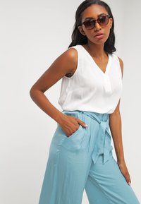Sleeveless white top with a v-neck and a single front button, paired with high-waisted light blue pants featuring side pockets and a tie waist.