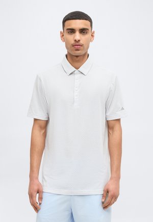 Ung mand med kort hår iført en lysegrå kortærmet poloshirt og lyseblå shorts, stående foran en ensfarvet baggrund.