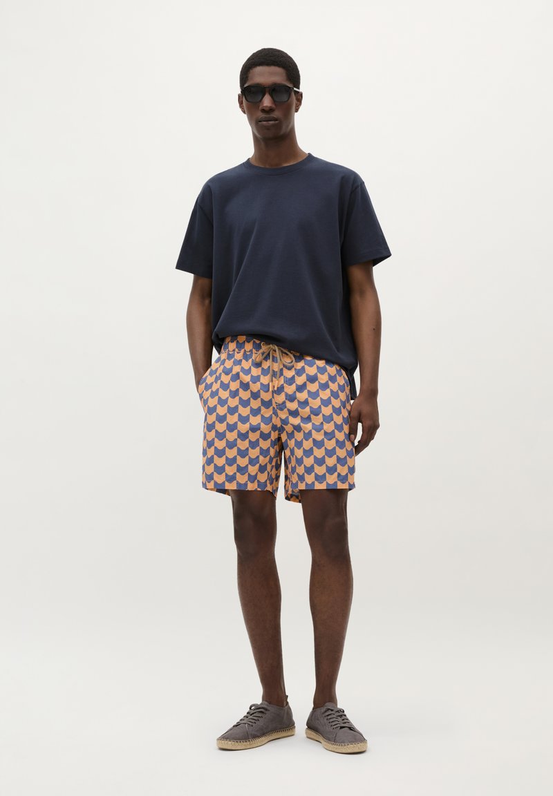 T-shirt en coton marine, short de bain orange et bleu à motifs avec taille élastique, et chaussures grises à lacets. Modèle portant des lunettes de soleil.