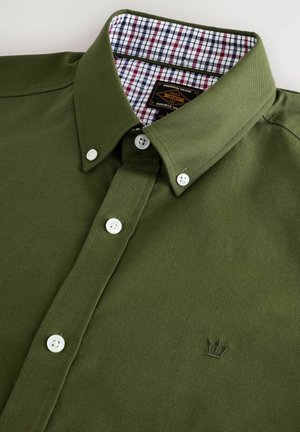 Zöld gombos ing hegyes gallérral, fehér gombokkal és kockás mintával a gallér belső részén, amelyen hímzett logó található a mellrészen.