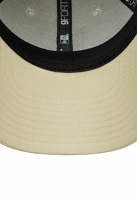 Base de una gorra New Era 9FORTY en beige y negro, mostrando la costura y la etiqueta de la marca dentro de la corona.