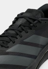 Chaussure de sport noire avec une tige texturée, des accents gris foncé, un design étroit et plusieurs bandes ton sur ton sur les côtés.