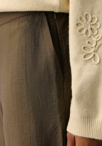 Jasno beżowy sweter z dzianiny z prążkowanymi mankietami, z haftowanym wzorem kwiatowym. W zestawie z oliwkowymi, teksturowanymi spodniami o szerokich nogawkach.