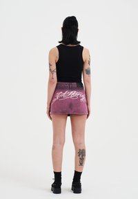 Denim minirok in vervaagd roze met witte geborduurde tekst op de achterkant. Model draagt een zwarte tanktop en chunky zwarte schoenen.