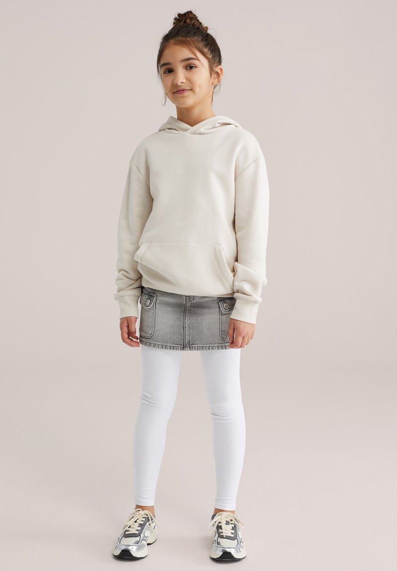 Világos bézs kapucnis pulóver első zsebbel, szürke farmer szoknyával zsebekkel, fehér leggingsszel és színes sportcipőkkel párosítva.