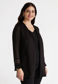 Blazer nero in tessuto liscio con maniche lunghe in pizzo trasparente. Design aperto davanti, senza chiusure e con tasche laterali.