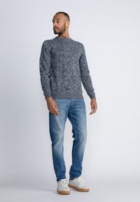 Marineblauwe gebreide trui met een ronde hals, gecombineerd met lichtblauwe jeans en witte sneakers met een rubberen zool. Model staat in profiel.