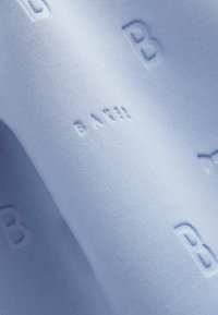 Svetle modrá tkanina s reliéfním nápisem obsahujícím písmeno "B" a slovo "BACH." Textura vypadá hladce a jemně.