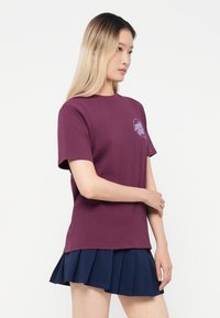 Maronnbrodt bomull t-skjorte med rund hals og korte ermer, med et lite grafisk motiv på brystet. Kombinert med en marineblå plisséskjørt.