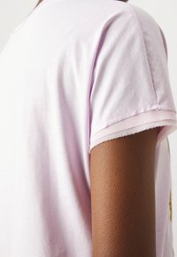 Ljust rosa kortärmad t-shirt med mjuk textur och rå kantdetalj vid ärmsluten, visad mot hudton.