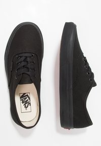 Zapatillas de lona negras con suelas de goma, que cuentan con seis ojales, cordones planos y una plantilla blanca con marca. Textura suave, diseño clásico de corte bajo.