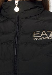 Czarna puchowa kurtka z wysokim kołnierzem, zamkiem błyskawicznym z przodu i subtelnym logo EA7. Posiada pikowaną fakturę oraz metalowy suwak.