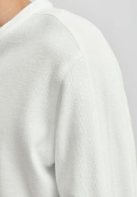 Primer plano de una tela de punto blanca y texturizada sobre el hombro y la parte superior del brazo de una persona, mostrando los detalles de la costura y suaves pliegues.