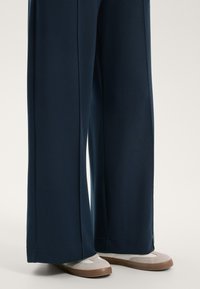 Morske široke hlače iz mehkega materiala, z ravnim krojem in robom, v kombinaciji s svetlimi teniskami v bež in beli barvi.