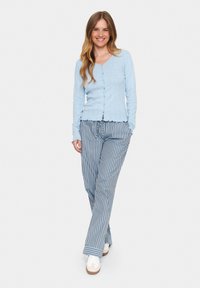 Světle modrý, žebrovaný kardigan na knoflíky s dlouhými rukávy, kombinovaný s volnými kalhotami modro-bílé pruhované barvy. Oblečení doplňují bílé tenisky.