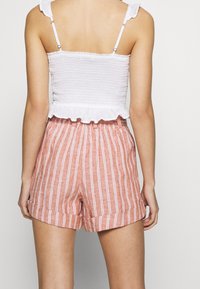 Vrouw in witte, rimpelige, mouwloze top en hooggewaide oranje shorts met witte verticale strepen, gezien vanuit de rug.