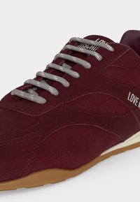 Gros plan d'une basket bordeaux avec des lacets gris, une semelle couleur sable et un texte blanc avec l'inscription "LOVE MOSCHINO" sur le côté et la languette.