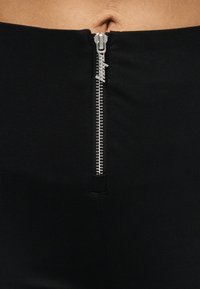 Κοντινό πλάνο ενός ασημένιου φερμουάρ σε μαύρο παντελόνι με ψηλή μέση, μερικώς ανοιχτό, αποκαλύπτοντας μια περιοχή γυμνού δέρματος πάνω από τη μέση.