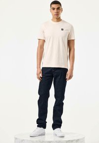 Tricou din bumbac bej deschis cu un patch negru pe piept, asortat cu blugi denim de culoare albastru închis și adidași albi, stând pe o suprafață de marmură.