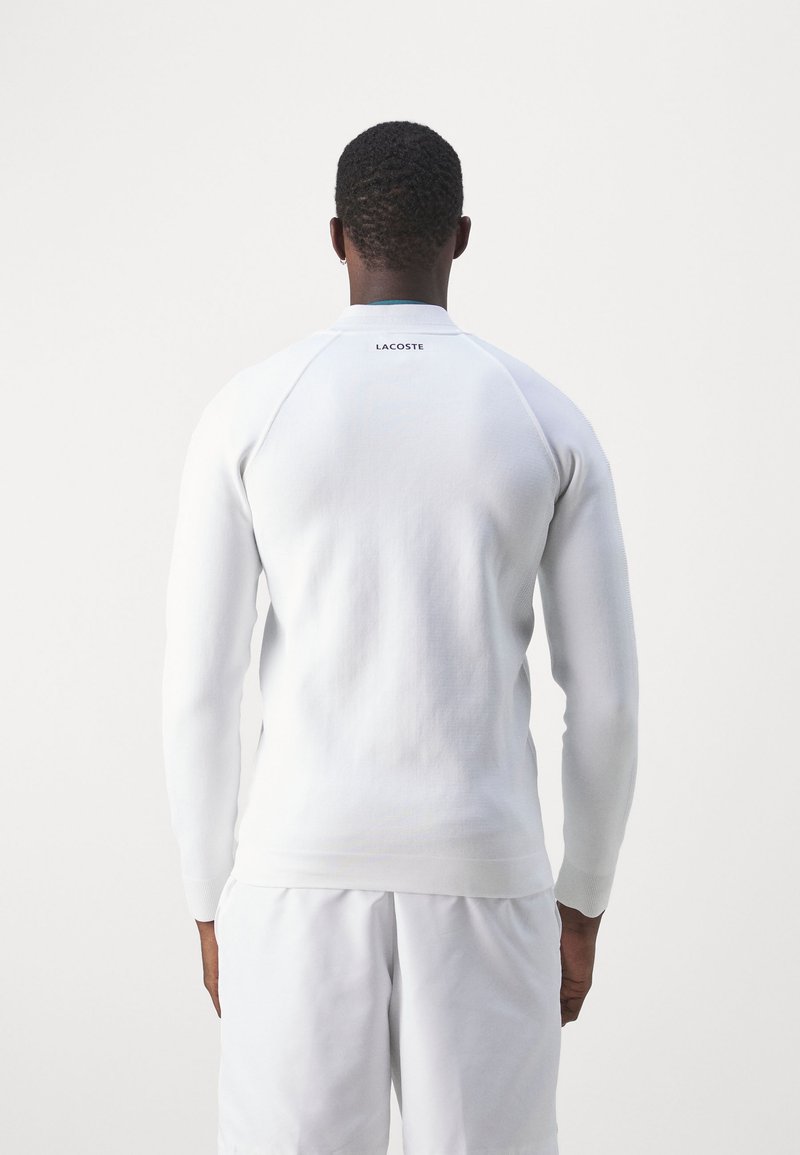 Sweatshirt blanc avec une finition texturée, des poignets et un ourlet côtelés, et un logo Lacoste discret au dos. Associé à un short blanc assorti.