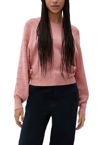 Femme portant un pull en maille rose avec des manches ballon texturées et un pantalon noir, debout devant un fond blanc.