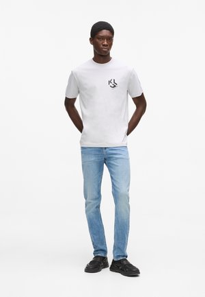 Jeune homme debout, mains derrière le dos, portant un t-shirt blanc avec un petit logo, un jean bleu clair et des chaussures noires sur fond blanc.
