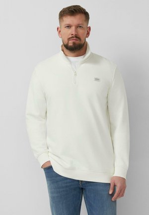 Mężczyzna ubrany w biały sweter z krótkim zamkiem błyskawicznym z naszywką na piersi i niebieskie dżinsy, stojący na tle jednolitego jasnoszarego tła.