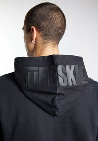 Zwarte hoodie met een gestructureerd, matte logo "TUFF SKULL" op de achterkant. Zachte stof met een ontspannen pasvorm en gedetailleerde stiksels.