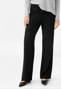 Svarta, vida byxor tillverkade av slät tyg, med en framficka och en subtil logodetalj. Matchas med spetsiga svarta skor.