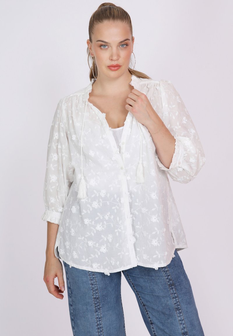 Weiße, durchsichtige Bluse mit floraler Stickerei, Knopfleiste, Kordelzug mit Quasten und drei Viertel-Ärmeln. Kombiniert mit blauen, ausgestellten Jeans.