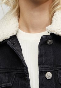 Veste en jean avec un col en shearling crème, tissu foncé et détails de boutons argentés ; superposée sur un pull en côte blanc.