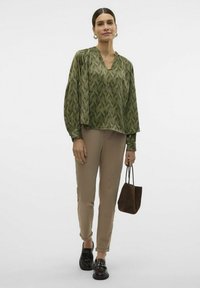 Zelená blouse s vzorem, s V-výstřihem, volným střihem a balónovými rukávy; spárována s béžovými kalhotami a černými mokasínami. Součástí je hnědá kabelka přes rameno.