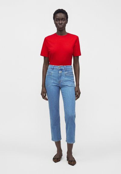 Femme aux cheveux courts portant un haut rouge à manches courtes, un jean court taille haute bleu clair et des ballerines à imprimé léopard, debout devant un fond blanc.