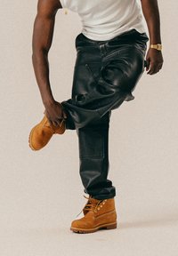 Svarte skinnbukser og brune suedesko med snøring, som fremhever et robust design. Buksene har en avslappet passform med lommer.